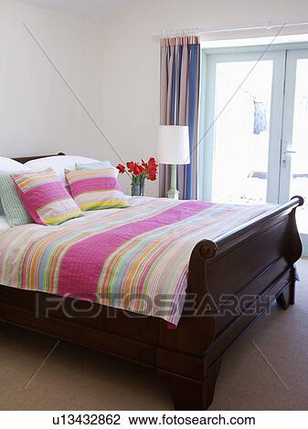 Stock Image - Colourful pink striped bedcover and cushions on mahogany lit bateau in modern white coastal bedroom. Fotosearch