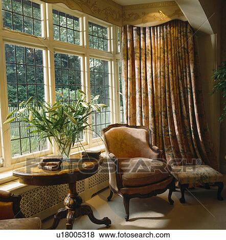 Comfy armchair and antique table in front of leaded window with opulent curtains in country living room View Large Photo Image Stock Photo - Comfy armchair and antique table in front of leaded window with opulent curtains in country living room. Fotosearch