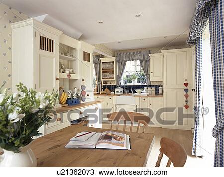 Stock Image - Cream fitted units in country kitchen with wooden table and chairs. Fotosearch
