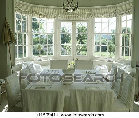 Stock Image - Cream linen loosecovers on chairs in cream country dining room with linen tablecloth. Fotosearch