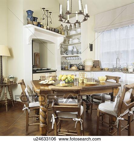 Stock Photograph - Cream striped cushions on antique chapel chairs in cream country kitchen. Fotosearch