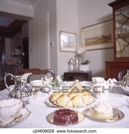Stock Photo - Dining room with silver jug and plate of scones on table set for tea with antique china and white tablecloth. Fotosearch