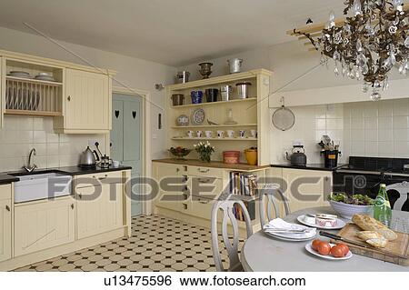 Stock Photograph - Georgian crystal chandelier above Victorian-style table and chairs in kitchen with cream units and chequerboard flooring. Fotosearch