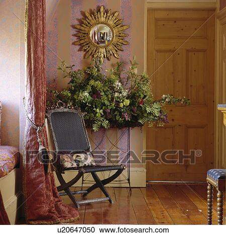 Gold sunburst mirror above floral arrangement on metal table in small hall with antique chair and mauve wallpaper View Large Photo Image Stock Image - Gold sunburst mirror above floral arrangement on metal table in small hall with antique chair and mauve wallpaper. Fotosearch
