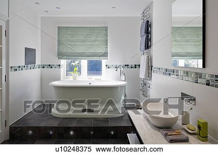 Stock Photography - Green blind on window above bath on split-level floor in modern bathroom with grey tiled floor. Fotosearch