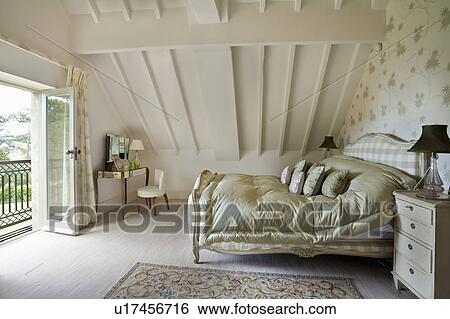 Stock Photograph - Green silk quilt on upholstered bed in cream attic bedroom with green floral wallpaper. Fotosearch