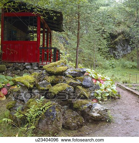 Stock Photograph - Large moss-covered rocks below red summerhouse in rainy country garden. Fotosearch