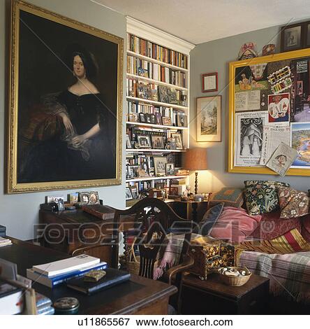 Stock Photo - Large oil portrait beside fitted bookshelves in traditional living room with poster-board above sofa piled with cushions. Fotosearch