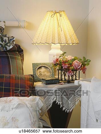 Stock Photo - Lighted lamp on bedside table with small vase of pink roses and fuchsias. Fotosearch