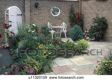 Stock Photo - Low-growing shrubs in paved townhouse courtyard garden with white metal table and chairs. Fotosearch