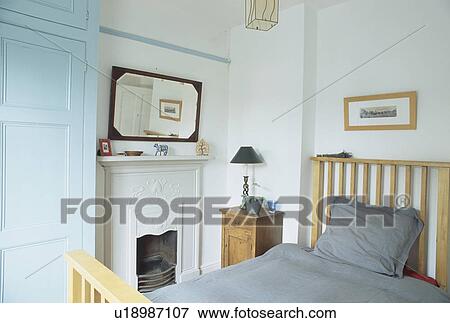 Mirror above fireplace in small white bedroom with grey bedlinen on wooden bed View Large Photo Image Stock Photo - Mirror above fireplace in small white bedroom with grey bedlinen on wooden bed. Fotosearch