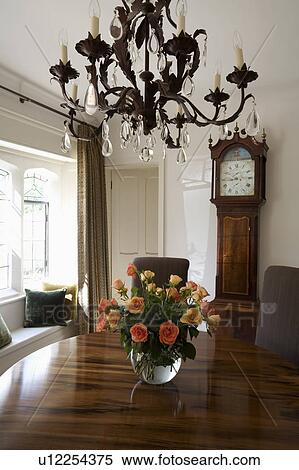 Ornate metal and glass chandelier above antique table with vase of orange roses in dining room with longcase clock View Large Photo Image Stock Photography - Ornate metal and glass chandelier above antique table with vase of orange roses in dining room with longcase clock. Fotosearch