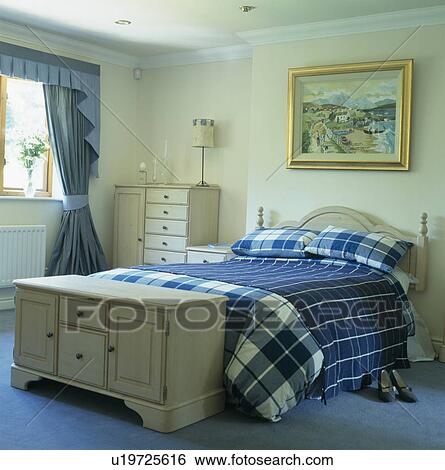 Stock Photograph - Pale wood chest below bed with blue checked bedcover in cream bedroom. Fotosearch