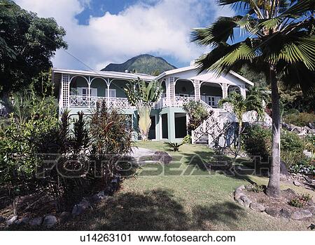 Stock Image - Palm trees and exotic shrubs in garden in front of traditional Caribbean house with balconies and veranda. Fotosearch