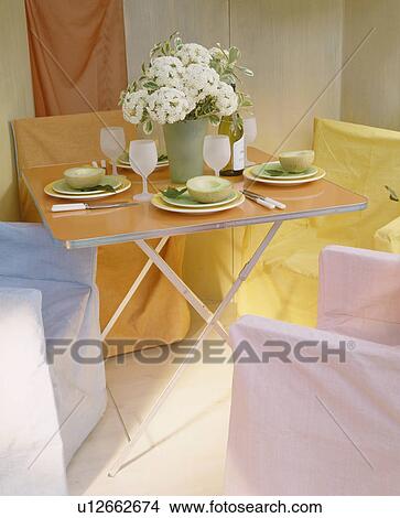 Picture - Pastel loose-covers on chairs around folding table set for lunch in economy-style dining room. Fotosearch
