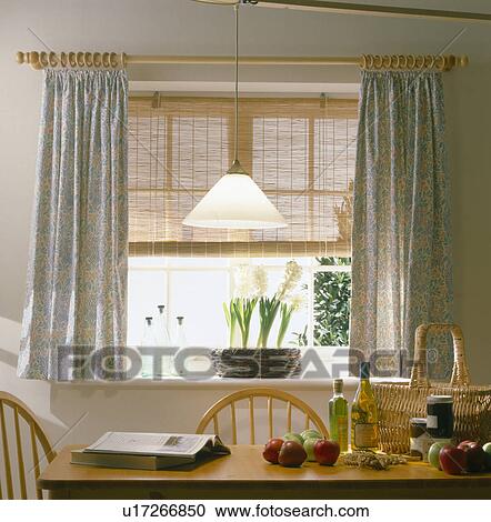 Stock Image - Pendant light above simple wooden table in economy-style dining room with floral curtains and cane blind at window. Fotosearch