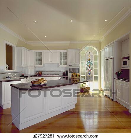 Stock Image - Polished wooden flooring in modern white kitchen with granite-topped island unit. Fotosearch