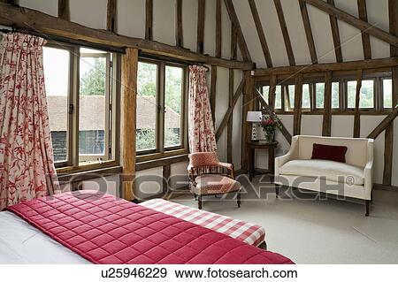 Stock Photo - Red quilt on bed in beamed country bedroom with small white sofa and red floral curtains. Fotosearch
