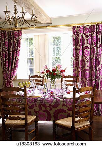Stock Image - Rush-seated chairs at table set for lunch in country dining room with pink cut-velvet Designer's Guild curtains and cloth. Fotosearch