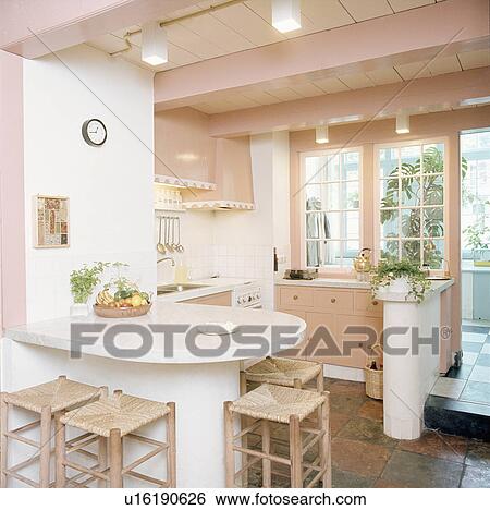 Stock Photograph - Rush-seated wooden stools at breakfast bar in pastel pink seventies kitchen. Fotosearch