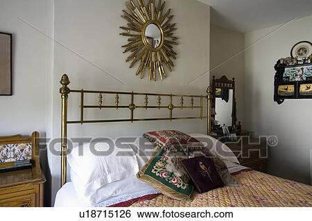 Stock Photograph - Starburst mirror above antique brass bed with white pillows and tapestry cushions in traditional bedroom. Fotosearch
