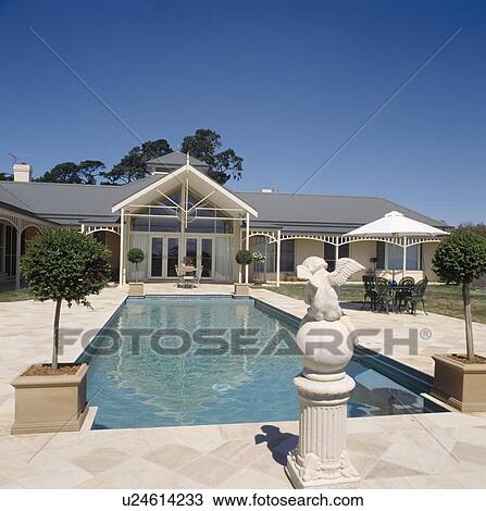 Stock Image - Statue on plinth on paved area surrounding swimming pool in front of single storey traditional Australian house. Fotosearch