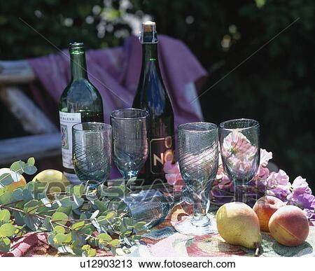 Stock Image - Still-Life of bottles of wine and wine-glasses on garden table with foliage and fruit. Fotosearch