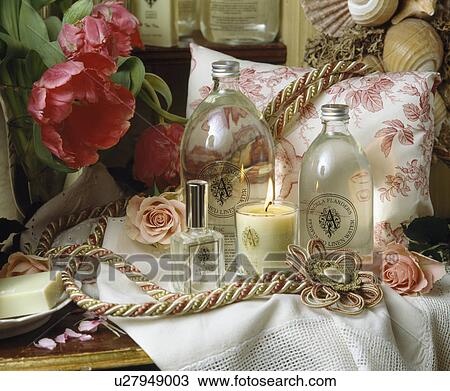 Stock Image - Still-Life of Victorian-style tableau with lighted candle and glass bottles on table with pink roses and floral cushion. Fotosearch