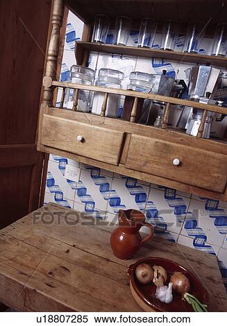 Stock Photography - Storage jars on pine rack above brown jug and bowl of vegetables on rustic wooden table. Fotosearch