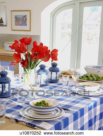 Stock Image - Table set for lunch with vase of red tulips and blue checked tablecloth with blue striped crockery. Fotosearch