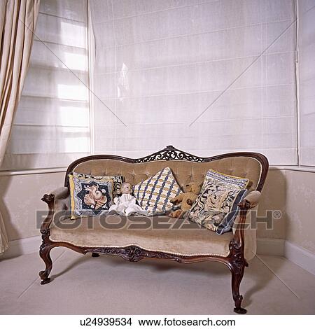 Picture - Tapestry cushions and vintage doll on beige velour upholstered antique sofa in front of window with white blinds. Fotosearch
