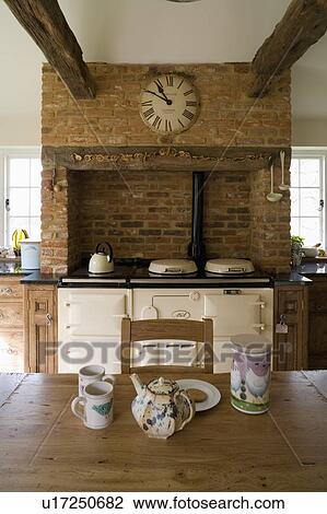 Stock Image - Teapot and cups on pine table in cottage kitchen with clock on Tudor brick wall above cream Aga. Fotosearch