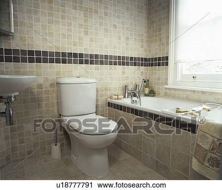 Stock Image - Toilet beside bath with tiled surround in modern beige tiled bathroom. Fotosearch