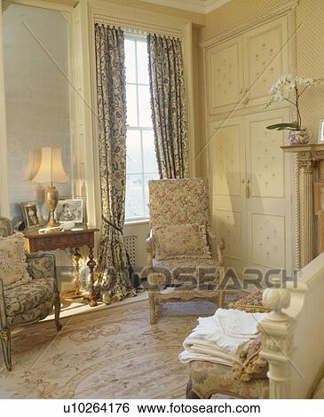Upholstered chair in front of fitted wardrobe and patterned blue curtains on window of country bedroom View Large Photo Image Stock Photograph - Upholstered chair in front of fitted wardrobe and patterned blue curtains on window of country bedroom. Fotosearch