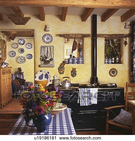 Stock Image - Vase of flowers and blue checked cloth on table in cottage kitchen with black Aga oven. Fotosearch