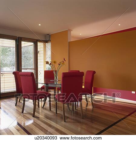 Stock Image - Vase of gerberas on glass table orange dining room with red upholstered chairs. Fotosearch