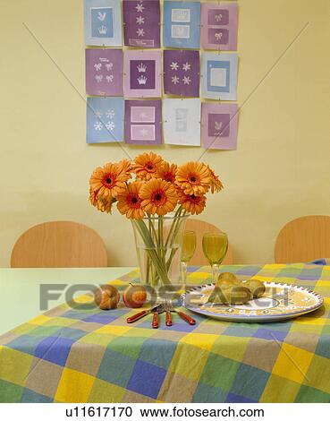 Stock Image - Vase of orange gerberas on dining table with yellow and green checked cloth. Fotosearch