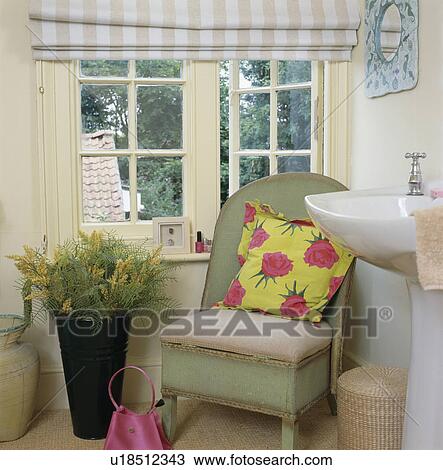 Stock Image - Vase of yellow mimosa beside green Lloyd Loom chair with yellow and pink cushion. Fotosearch