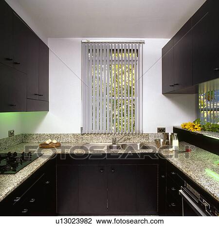 Stock Image - Vertical slatted blind on window above sink in white kitchen with black fitted cupboards and granite worktops. Fotosearch