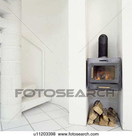 Stock Photo - Wall-mounted wood-burning stove above pile of logs in small white cottage hall with spiral staircase. Fotosearch