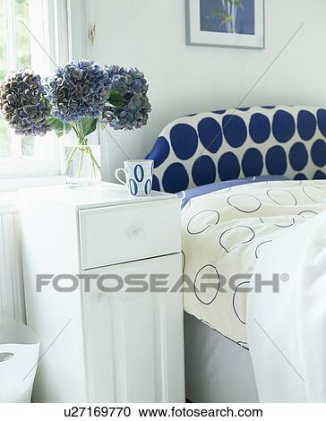 Stock Image - White bedside table beside bed with blue+white spotted headboard. Fotosearch