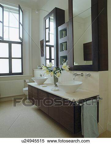 Stock Photography - White ceramic bowl-basins on vanity unit below mirrored cabinets in modern monochromatic bathroom. Fotosearch