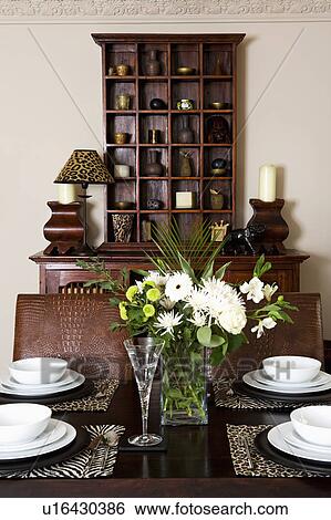 Stock Photograph - White crockery and animalprint placemats on table in dining room with dark wood shelf unit. Fotosearch