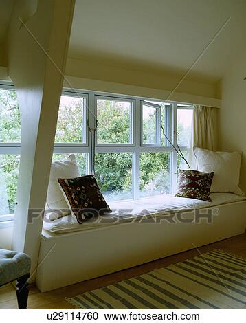 Stock Image - White cushions on window-seat below window in modern cottage living room. Fotosearch