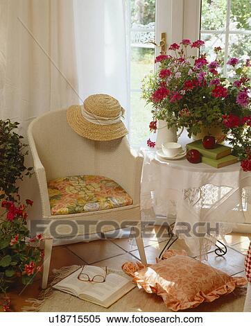 Stock Photography - White Lloyd Loom chair beside table with white cloth and pot of red geraniums. Fotosearch