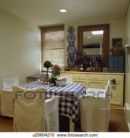 Stock Image - White loose-covers on chairs in dining room with checked cloth on table. Fotosearch