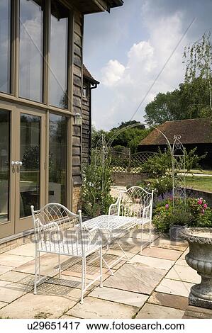 Stock Photo - White metal chairs and table on paved patio in front of barn conversion. Fotosearch