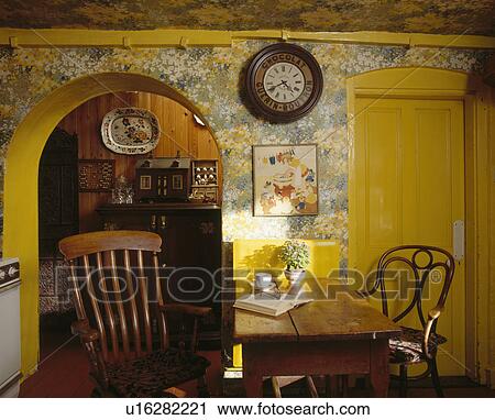 Stock Image - Windsor and Bentwood chairs at wooden table in dining room with floral wallpaper and yellow paintwork. Fotosearch