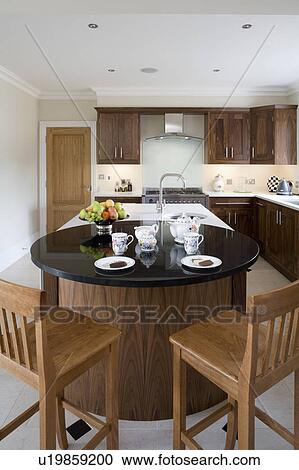 Stock Image - Wooden chairs at circular breakfast bar with cups and plates on black granite worktop in modern walnut kitchen. Fotosearch