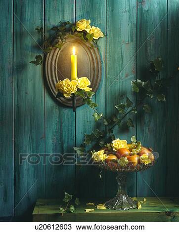 Stock Image - Yellow roses on mirror with lighted candle on green painted wooden wall above table with bowl of fruit and roses. Fotosearch